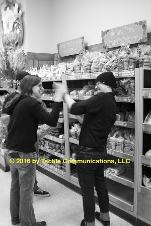 Image description: black and white (b&w) picture of three people, two females and one male, in Trader Joe’s (a food store). From left to right, a male, Ray, is slightly hidden behind Hayley on the left and aj is standing to the right. The three are standing in front of a shelf stacked with different types of bread. Hayley is tactiling with aj, both are smiling big. *Image © 2016 by Tactile Communications, LLC