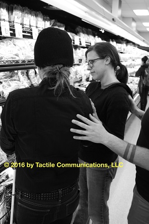 Image description: b&w picture aj and Hayley, in a refrigerated section, looking at a dairy product, while Ray stands behind them with his hands touching both. aj is wearing a black winter hat and black shirt. Hayley has a pair of glasses on, hair pulled back in a pony-tail, and is wearing a black shirt. *Image © 2016 by Tactile Communications, LLC