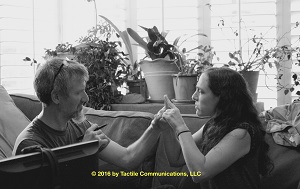 Image description: b&w picture of two people, a man (Roger) and a woman (Terra), sitting on a sofa, ProTactiling. Terra is sitting on the right, while Roger is sitting on the left. Terra is signing, while Roger is tactiling with one hand and the other is holding a pen. In the background, several different types of plants are sitting on a table. *Image © 2016 by Tactile Communications, LLC