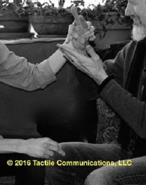 Image description: black and white photograph of two people sitting on a sofa, ProTactiling. The person on the right, Roger, is signing as the person on the left (face is not seen) is tactiling with one hand while the other hand is using PT, signing, "No" on Roger's leg.  *Image © 2016 Tactile Communications, LLC